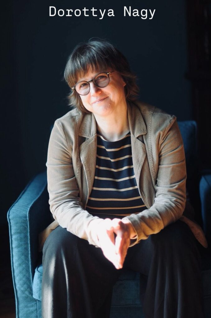 Professional headshot of a counsellor seated in a blue armchair against a dark background, smiling gently with a relaxed posture. She wears round glasses, a striped top, and a neutral-toned jacket, with soft natural lighting highlighting her face and hands. The image conveys a calm, approachable, and empathetic presence suitable for a therapy or counselling website.